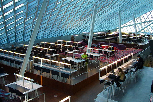 Interior of the Seattle Public Library. We see a sloping glass ceiling with a steel diamond pattern and great steel beams supporting the rood, also at an angle. There are rows of library books, people sitting at wooded tables, and lots of natural light.