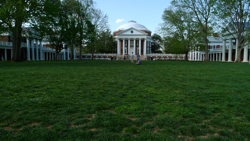 The Great Lawn, Rotunda, and Pavilions at the University of Virginia. The image shows a green lawn flanked by rows of pavilions made of red brick and white painted wood and stone. A large Neoclassical structure sits at the far end. It has a domed roof, columns, and a triangular pediment.