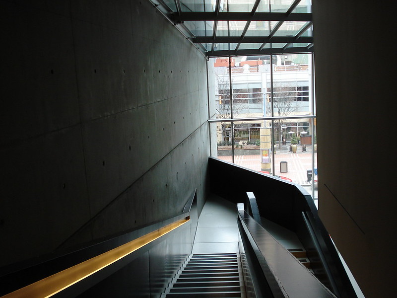 From the interior of the Contemporary Art Center, on the staircase, looking out. The walls and stairs are dark with the bright yellow railing. The city street is shown through floor to ceiling class windows.