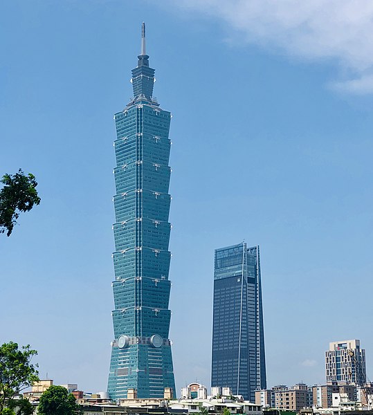 A tall skyscraper seen from a distance. The tower (Taipei 101) is a blue-green in color, made of glass, and moves upwards like blocks, rather than a smooth tower.