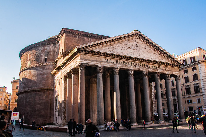 The ancient Roman Pantheon. A large structure with a triangular pediment, Corinthian columns, and a large drum base holding a massive dome. Made of concrete.