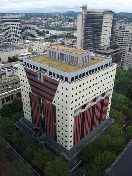 A colorful Postmodern building in Portland, OR. The colors on the structure are cream, teal, and a rusty red. There are green trees surrounding its base.