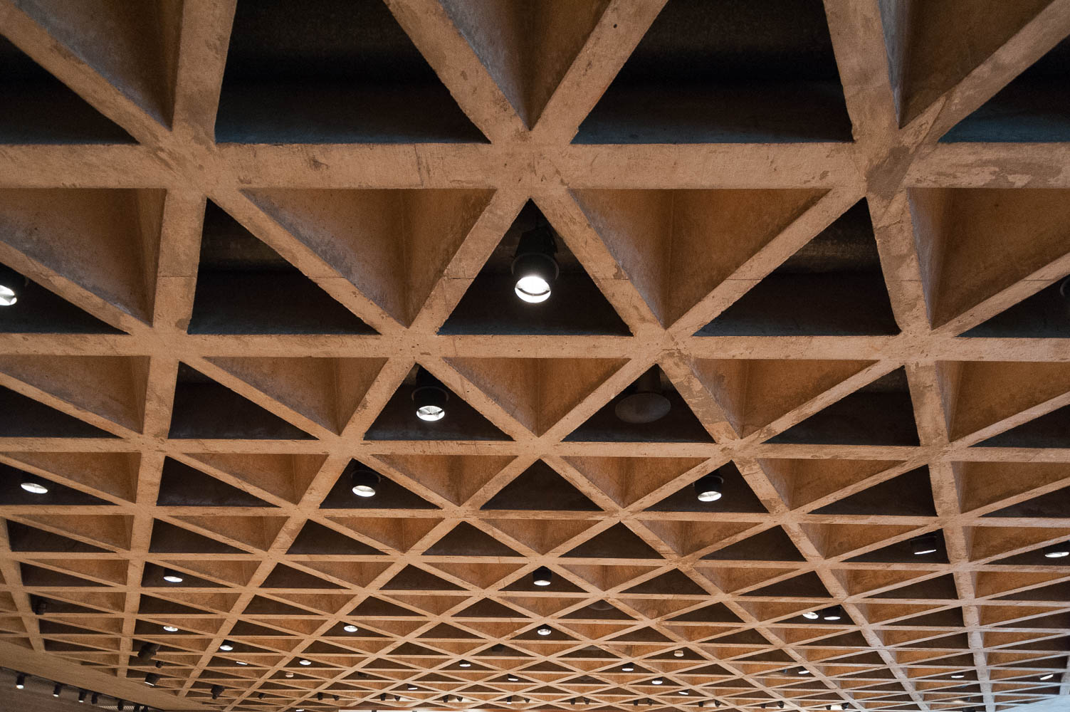 A view of a concrete ceiling with recessed track lighting. The ceiling is shaped as a series of tetrahedra.