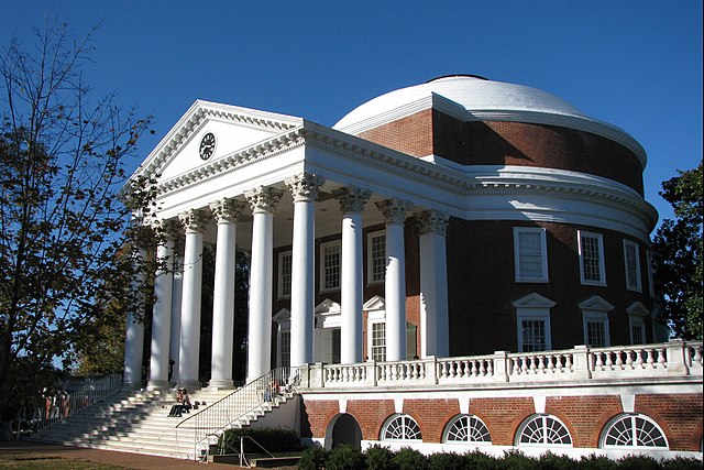 A Neoclassical structure, the Rotunda or library at the University of Virginia, seen from an angle. There are steps leading to a porch with tall, Corinthian columns holding up a triangular pediment with a round clock at its center. The side of the dome is visible, as is the round drum it rests on. The materials a red brick and white painted wood and stone.