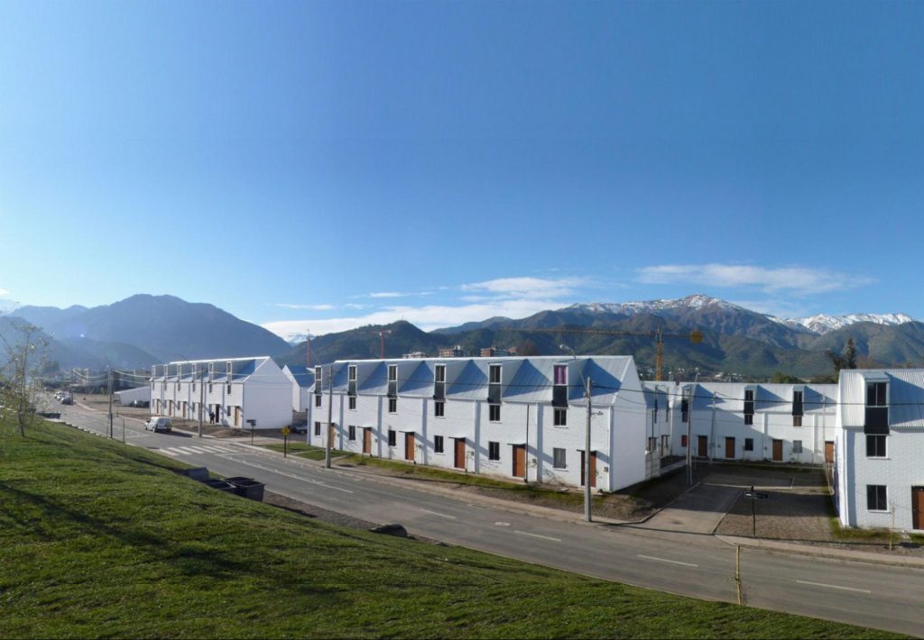 A series of houses with mountains in the background and a grassy landscape in front. The houses are white in color with blue-gray roofs.