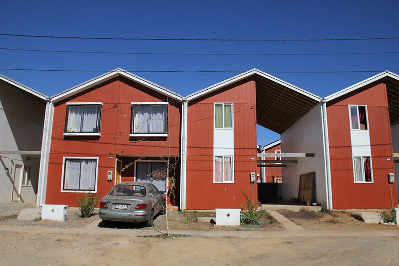 Image shows a row of houses. One has been finished by the owners, the other main one shown is half finished. They are rust red in color with light roofs and windows.