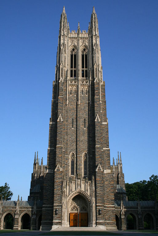 A Gothic Revival style church tower made of a dark stone with lighter stone elements highlighting the tops of the towers and the entrance portal.