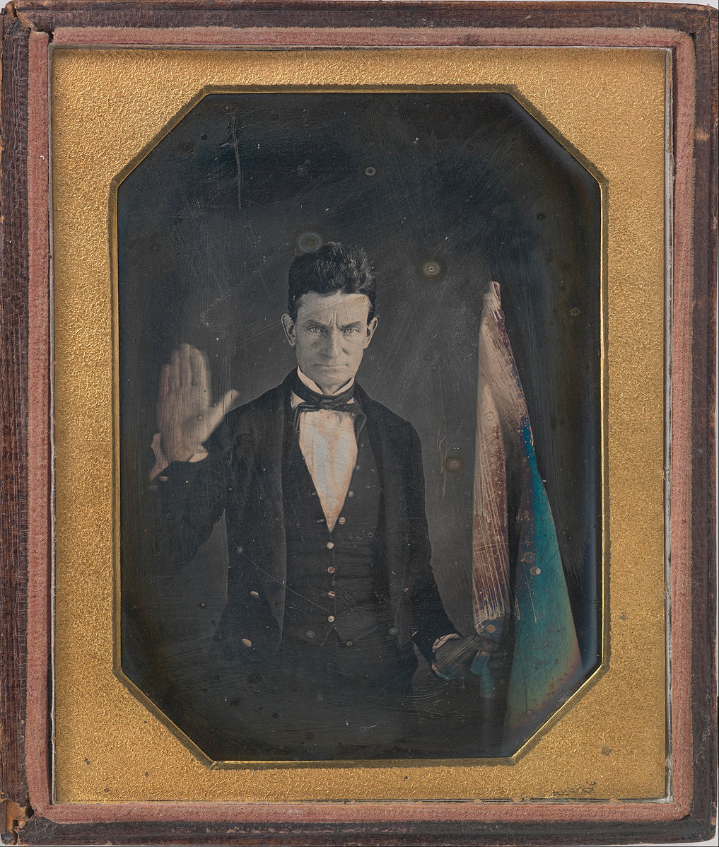 A framed portrait of John Brown wearing a tuxedo style suit. He has his right hand raised and holds a flag in his left hand. The flag has red and greenish blue coloring.