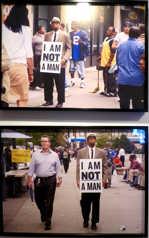 Two framed photographs vertical on a wall. Both show a Black man walking down a city sidewalk wearing a sign that says "I AM NOT A MAN" in all black capital letters on a white background..