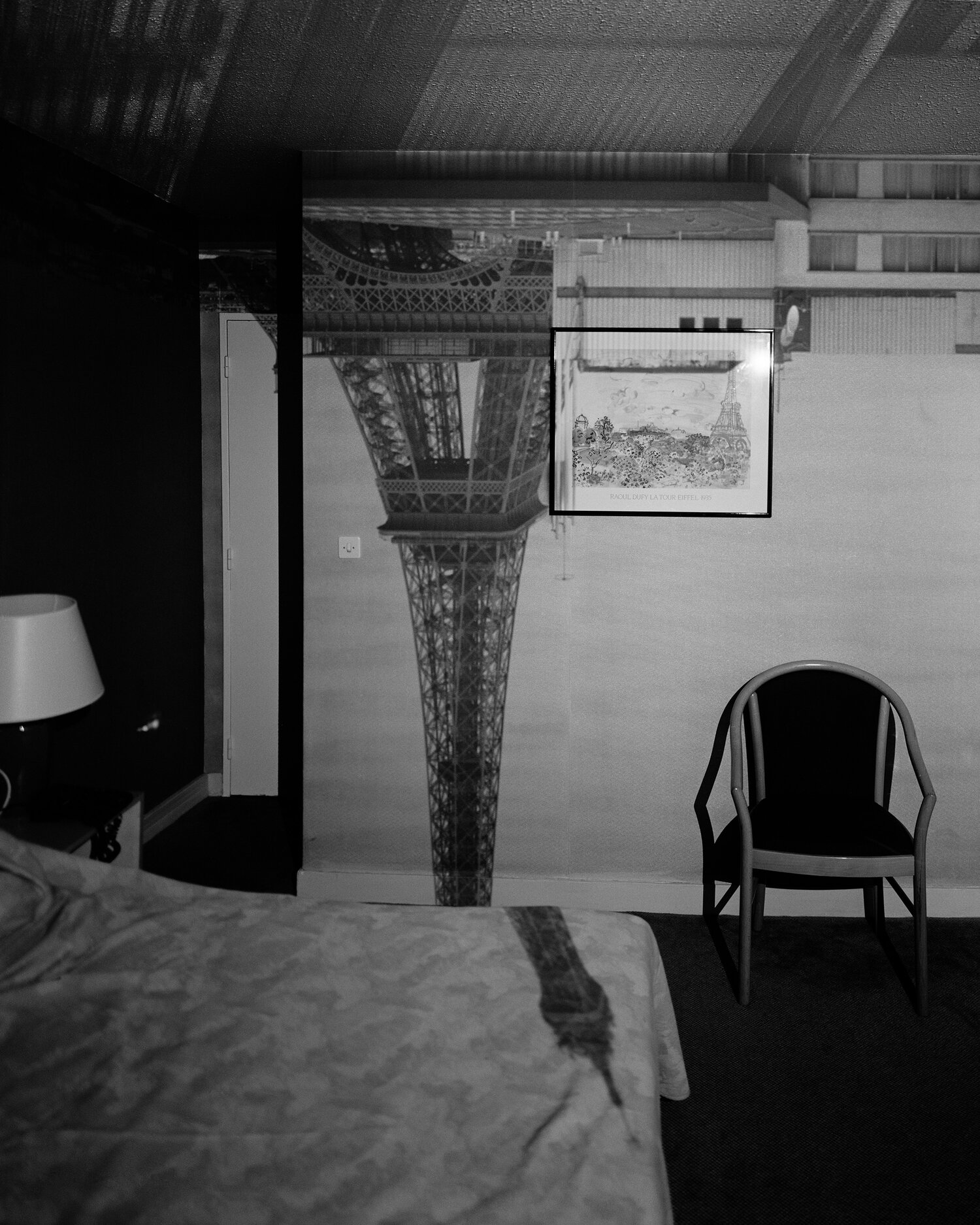 A black and white photograph of a hotel room with a bed and chair. A shadow of the Eiffel Tower is upside down on the back wall and across the bed.