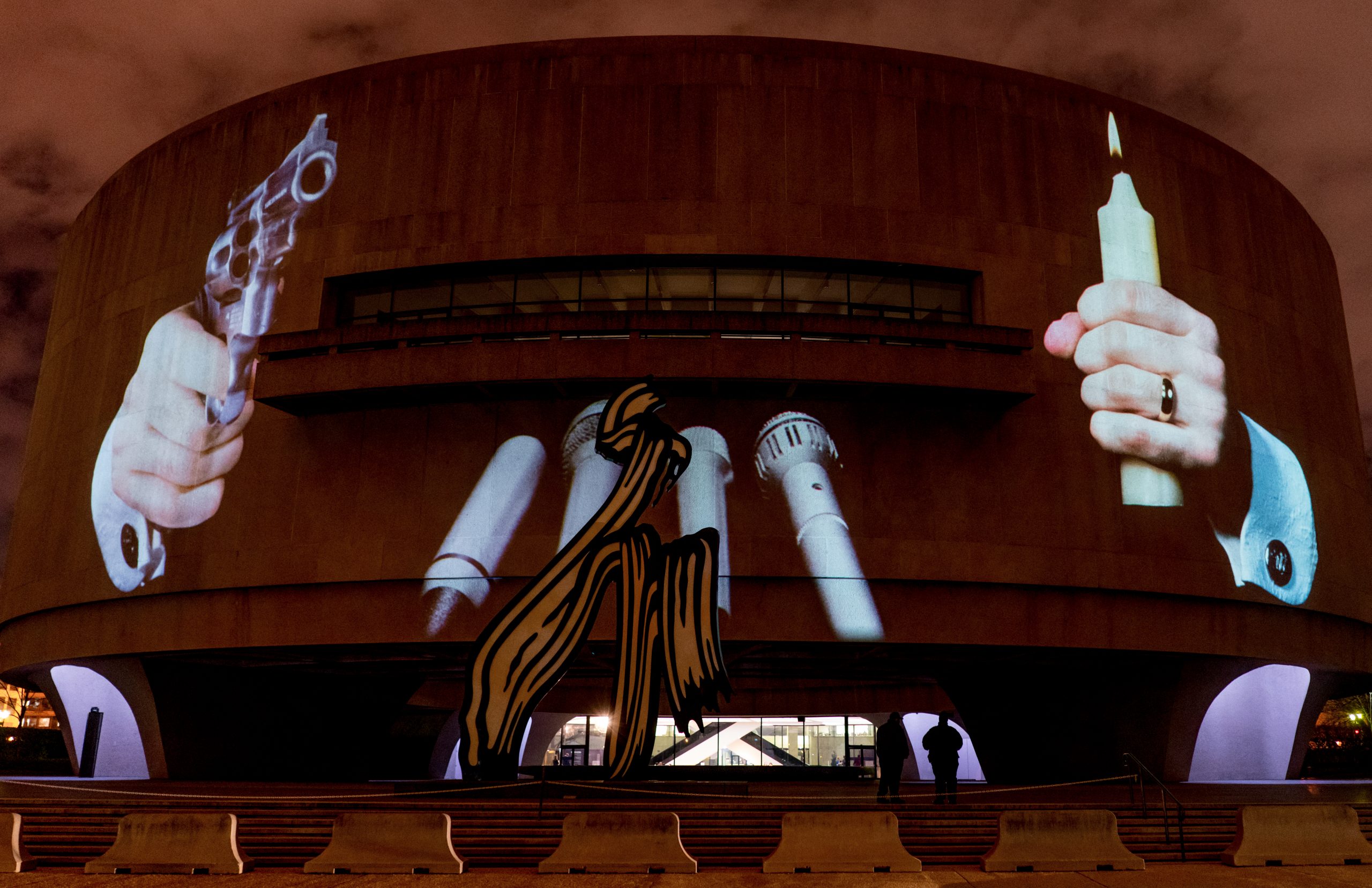 A photograph of the facade of the Hirshhorn Museum at night with a projection of a large scale hand holding a revolver, a hand holding a burning candle, and a bank of microphones in between.