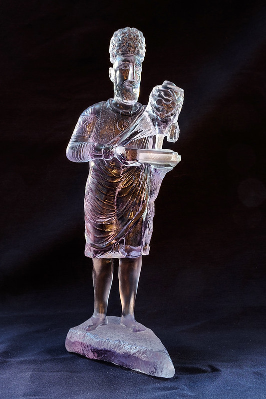 Translucent sculpture of a man standing on a rock with short curly hair and a draped tunic that lands above his knees. He carries an object in his arms.