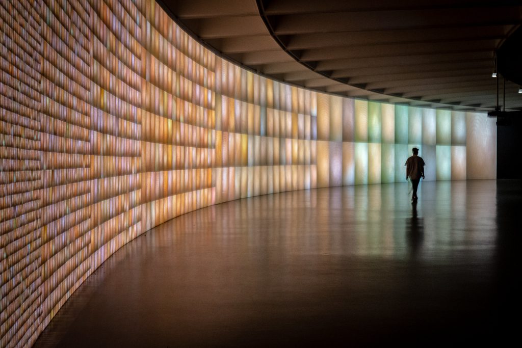 Person walking in front of a curved wall covered with patches of light in a variety of pastel colors.