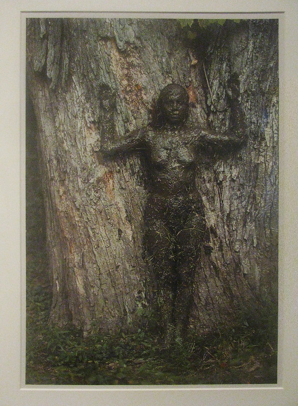 An upright female figure stands against a tree truck, completely covered in mud. The woman's arms are partially raised; her elbow and forearm creating a right angel.