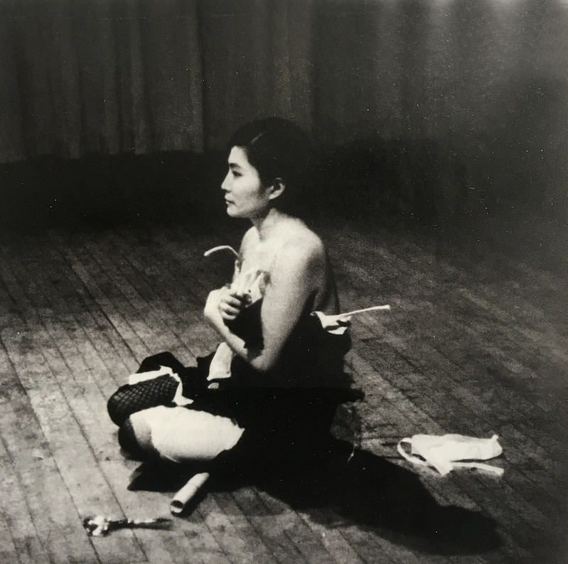 A woman, seen from the side, sits cross-legged on a stage. She holds her clothes up to her body. Bits of clothes that have been cut from her are seen behind her on the stage.