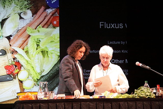 Two figures stand on a stage with the ingredients for a salad in front of them. The woman on the right, Alison Knowles, reads from a paper and behind the two figures is a screen with some words visible including &quot;Fluxus&quot; and a picture of a salad.
