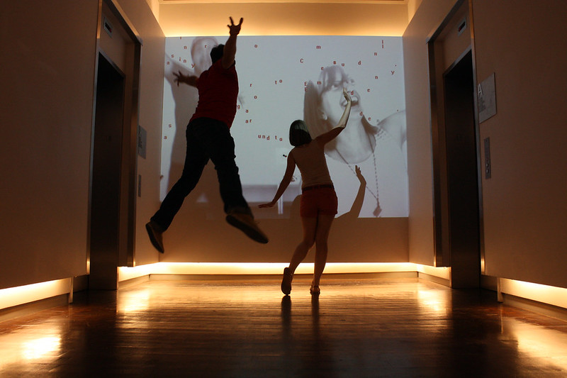 Image of two people, seen from the back, jumping. The figure on the left has their arms in the air and feet of the ground, while the figure on the right has both feet on the ground and arms partially raised. They both stand in front of a screen showing a digital projection of letters dropping down.