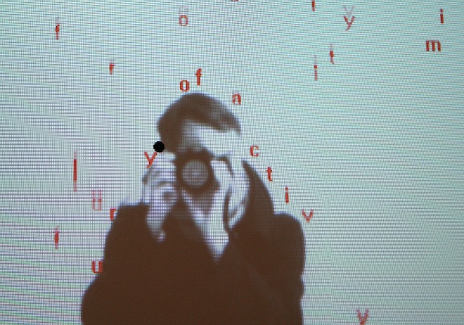 A figure, seen from the front, in front of a screen of Camille Utterback's &quot;Text Rain&quot;. Red letters appear to fall over the figure's shoulders/