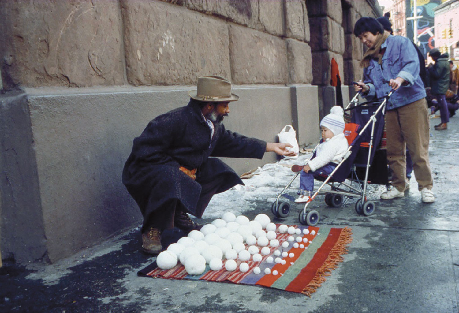 A man crouches down behind lines of snowballs arranged according to size. He passes one snowball to a baby wearing a white coat and hat, sitting in a stroller.