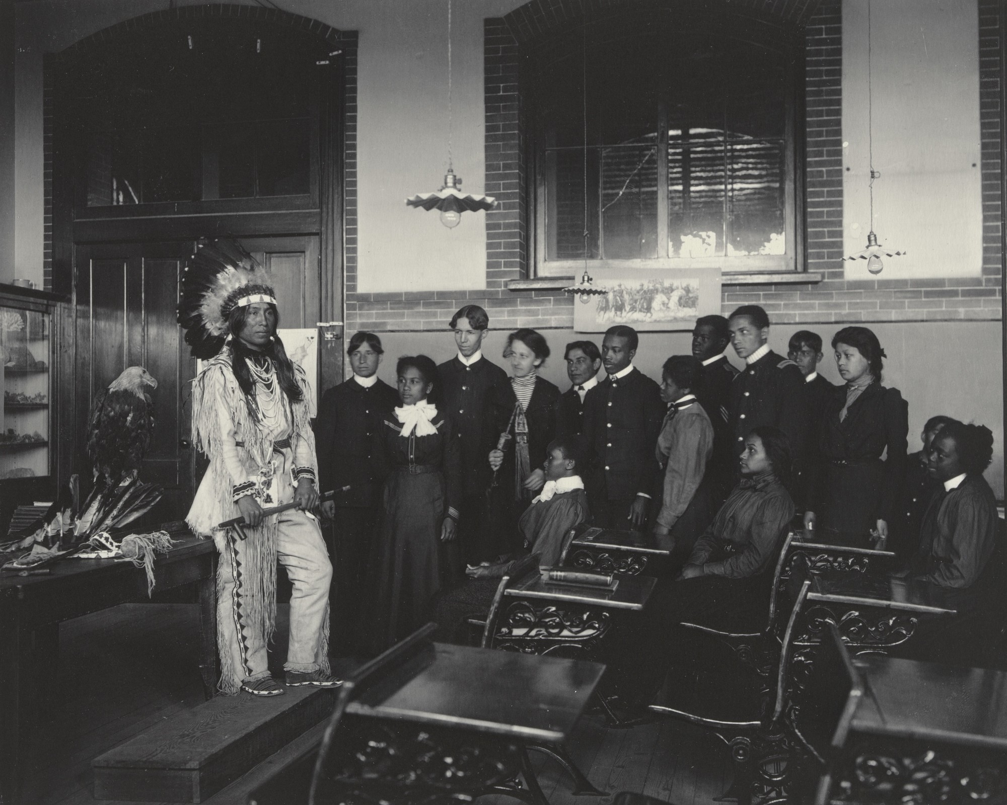 A black and white photograph of a group of students standing at the back of a classroom with desks arranged in front of them. They look at a man dressed in costume of a Native American. He is standing next to a taxidermied bald eagle.