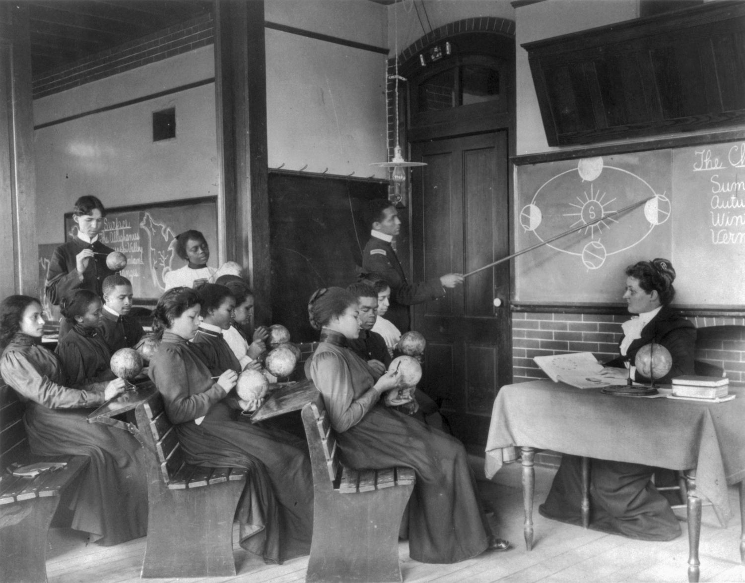 A black and white photograph of a classroom. The students are sitting on benches and hold small globes, looking at them and gesturing with pointers. A woman sits at the front of the room behind a desk. A man stands in the background and uses a long stick to point to a diagram on the board behind the woman.