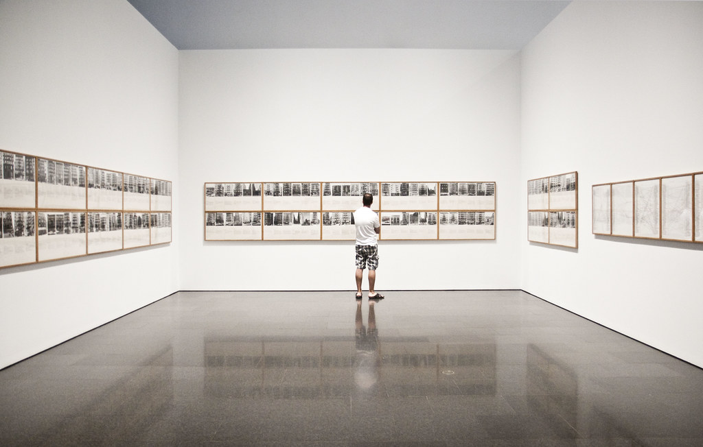 A photograph of a visitor viewing two long rows of photographs and text that are displayed on three white walls of a gallery.