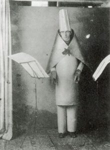 A black and white photograph of a man standing, dressed in a tall hat, cape, and lobster-like claws; the figure stands in front of a music stand with paper on it.