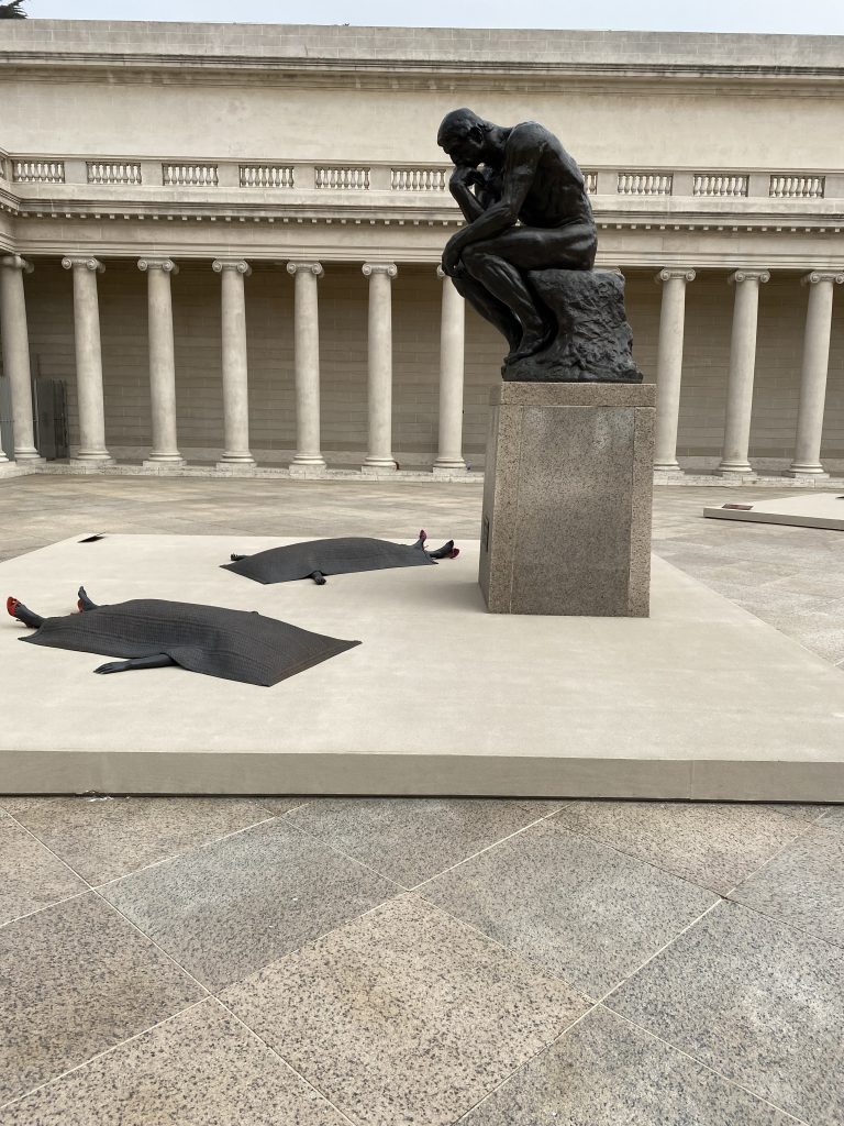 Image of Rodin's The Thinker, looking down at two sculptures by Mutu in the Legion of Honor Courtyard.