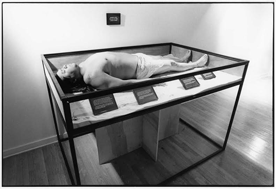 A black and white photograph of a person dressed in a loincloth lying in a museum display case with text labels.