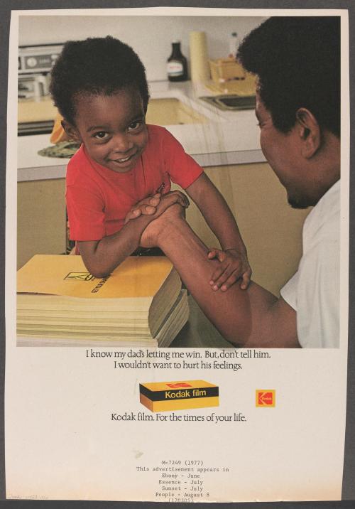 A photographic magazine advertisement for Kodak film depicting a boy in a kitchen arm wrestling his father. The boy looks into the camera smiling with his arm propped on a stack of telephone books. The man has his back to the camera and is croped on the right side. Text and an image of a box of camera film occupied the lower section of the page.