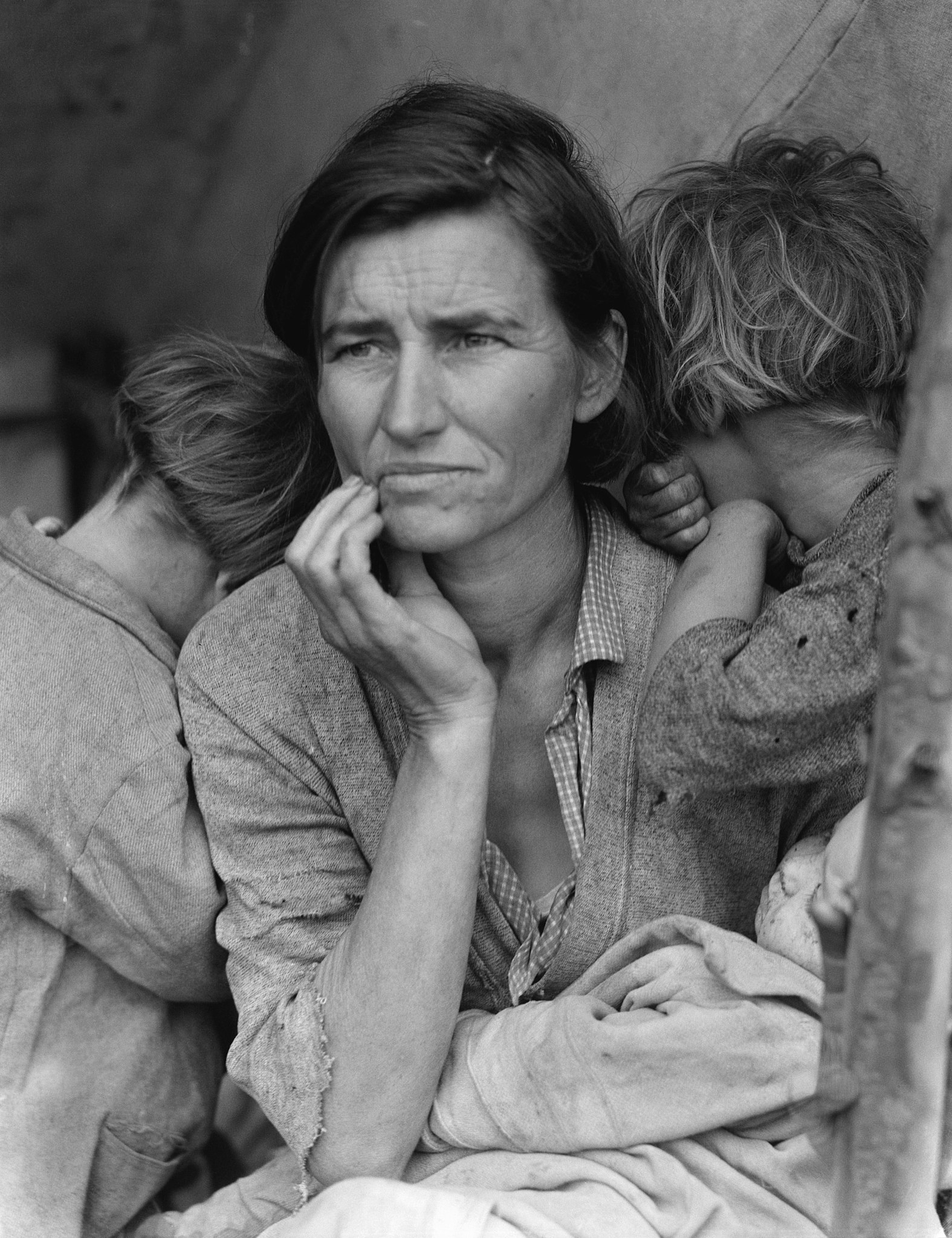 A black and white photograph of a woman seated. Her right hand touches her face near her chin. Two children bury their heads in her shoulder, one on each side of her. An infant is wrapped in a blanket on her lap.