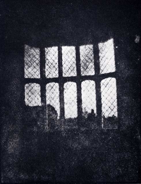 A black and white photograph looking out of a latticed window. The photograph is very dark except for the grid of bright white panes.