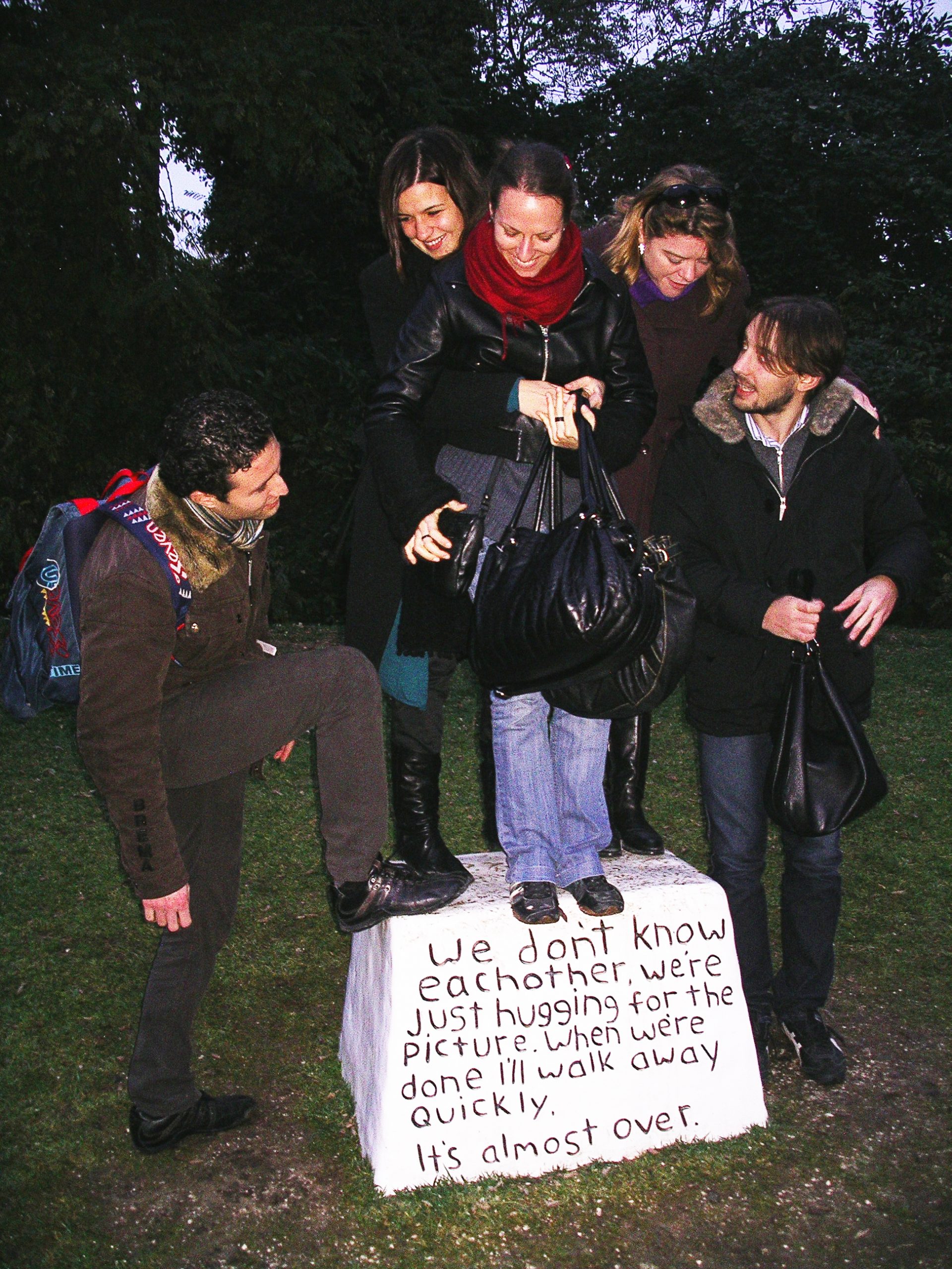 Five people attempting to stand on a pedestal inscribed with words.