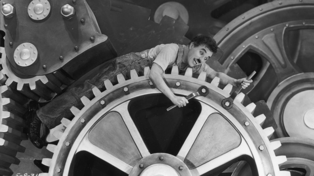 Charlie Chaplin caught in a gear, trying to fix it with wrenches.