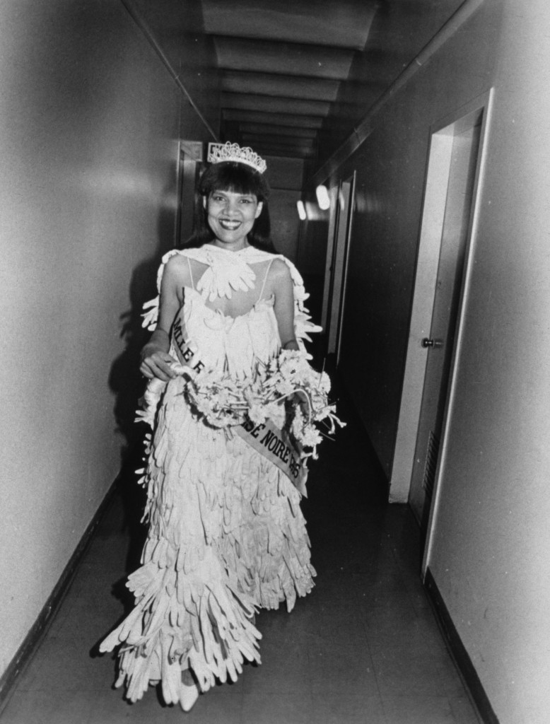 The artist Loraine O'Grady wearing a dress made of white gloves sewn together, holding a bouquet, looking directly at the camera and smiling. The photograph is black and white.