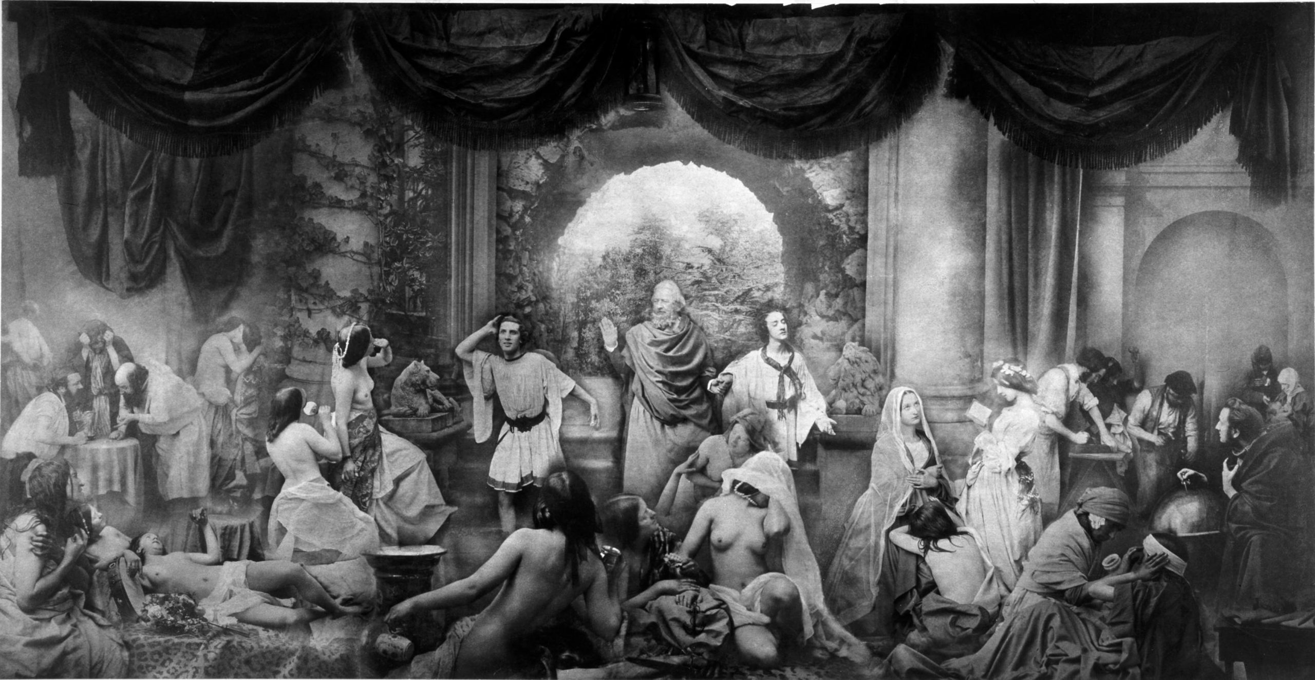 A long rectangular black and white photograph of small groups of people engaged in different activities from gambling to sewing. People are wearing classical drapery or are partially nude. Three men stand in the center in front of an arch. The central person raises his right hand. The two men on either side look and lean in opposite directions away from him.