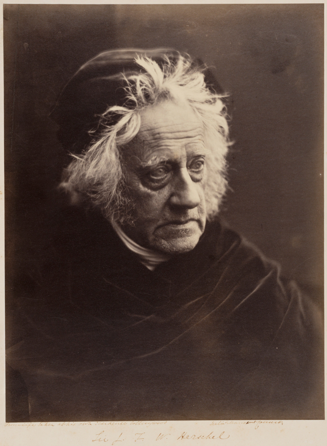 A black and white photograph of Sir John Herschel. He wears a cap, looks to his left, and is photographed from the chest up.