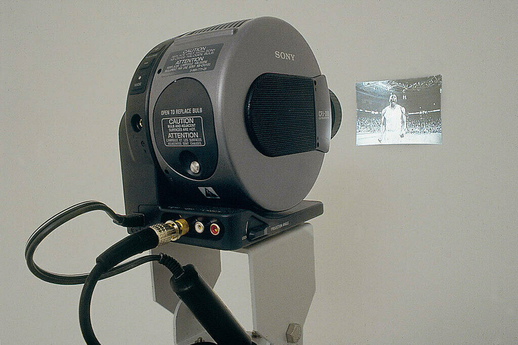 A projector, seen from the back, projects an image of a basketball player onto a blank, white wall.