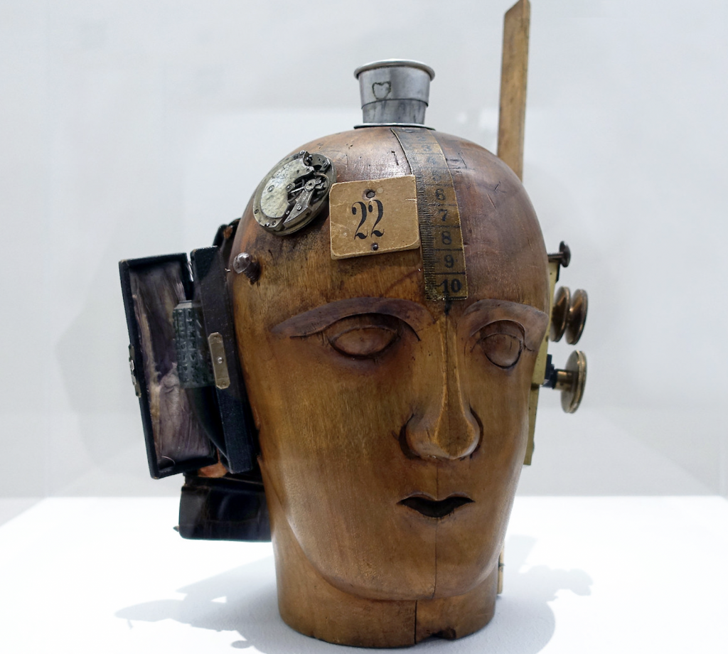 Dada sculpture of wooden head with rulers, and other mechanical objects attached to it.