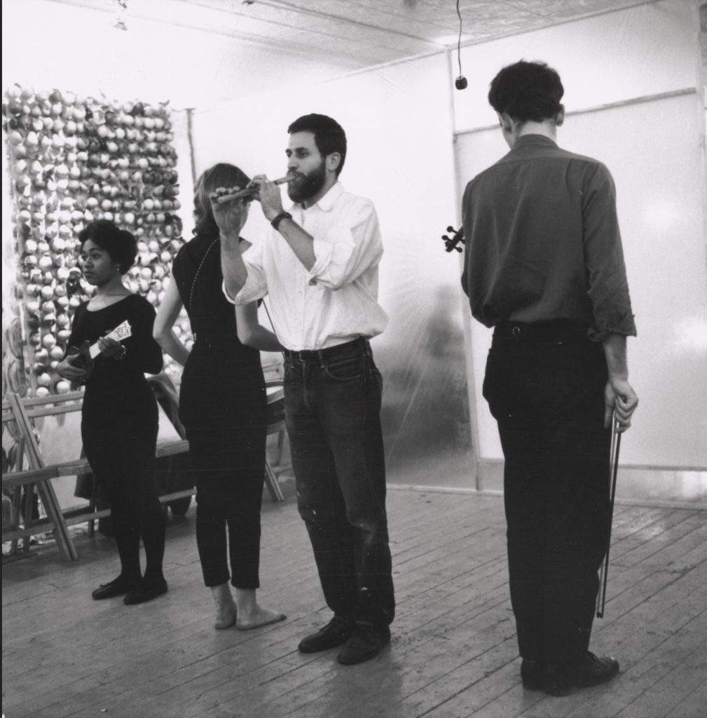 Black and white photo of four people standing in a room, two with their backs to the camera, one performing on a flute.