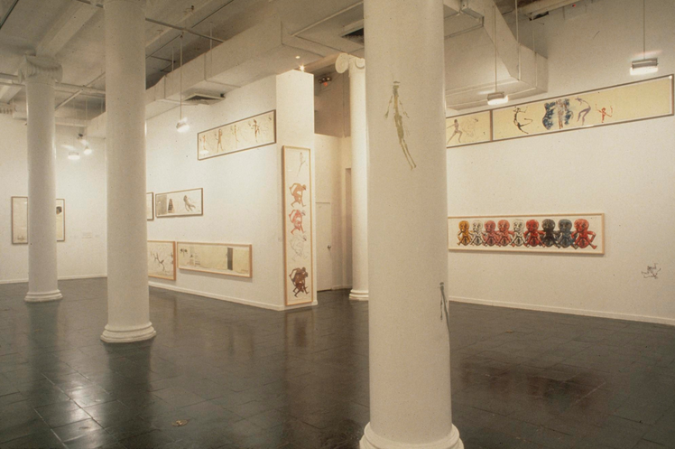 A photograph of the interior of a gallery with a row of columns in the foreground and prints hanging at different levels on the walls behind.