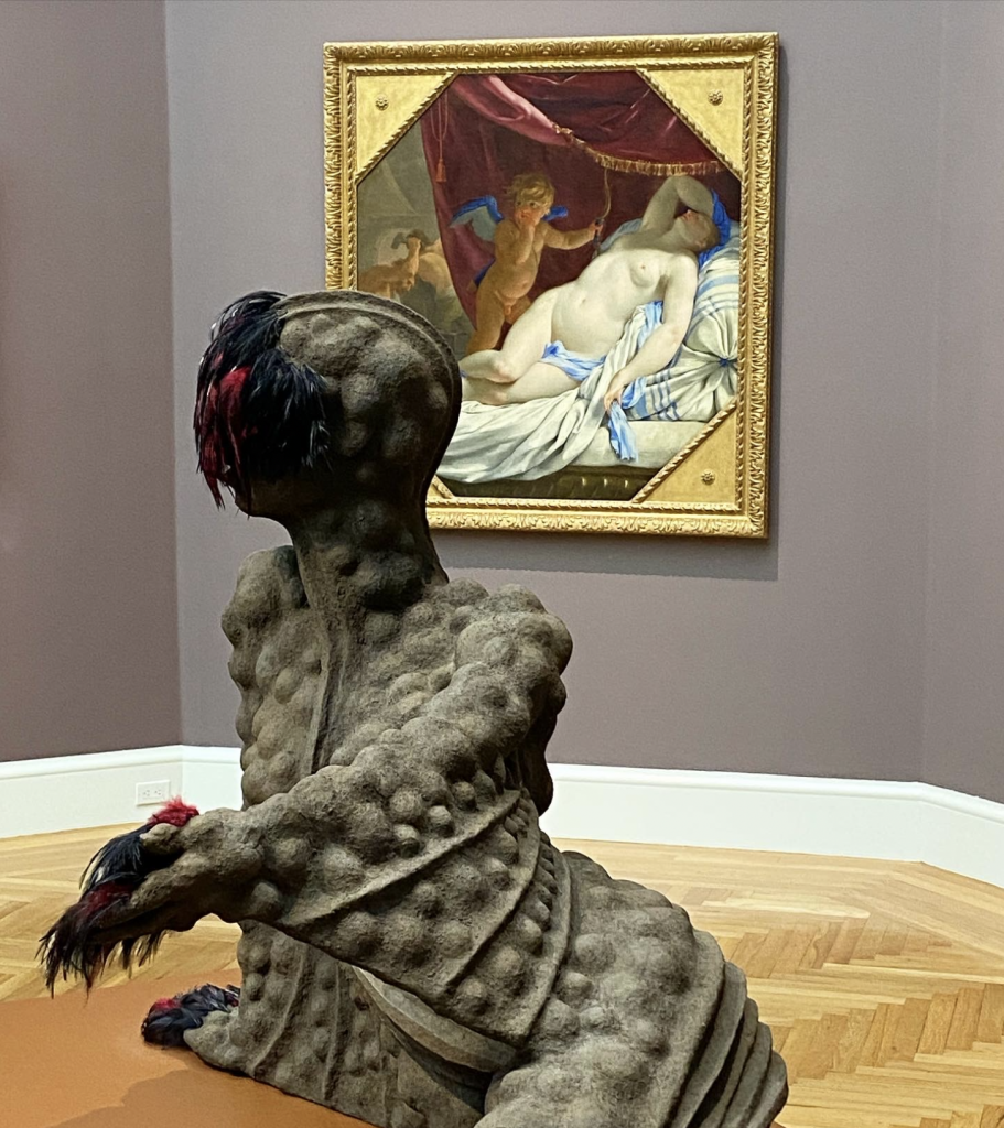 An afro-futurist sculpture by Wangechi Mutu with a European painting of a naked white woman behind it.