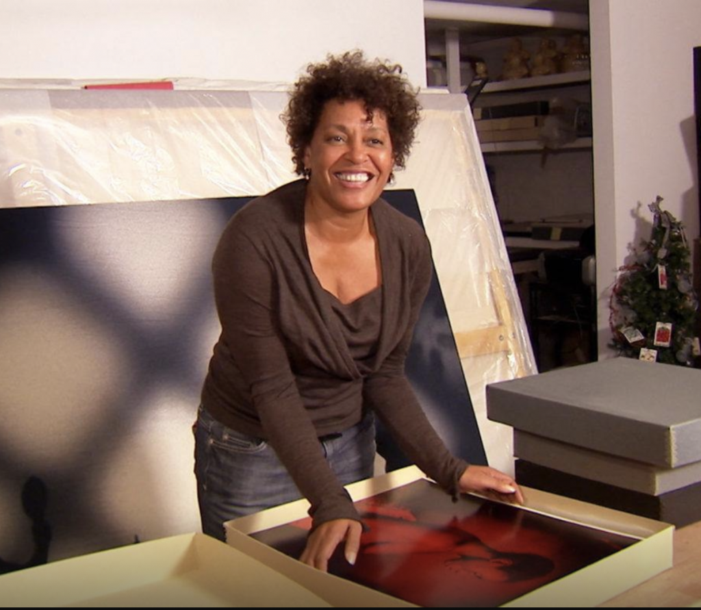 A smiling woman, the photographer Carrie Mae Weems in her studio, about to pull a photograph out of file.