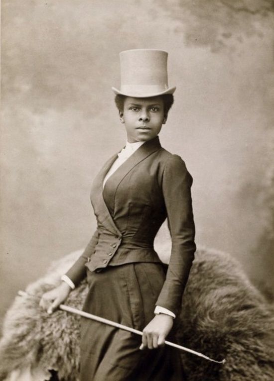 A black and white photograph with a warm undertone. A portrait of a woman equestrian turned facing the viewer in three-quarter view. She wears a riding uniform, a top hat, and holds a riding crop.