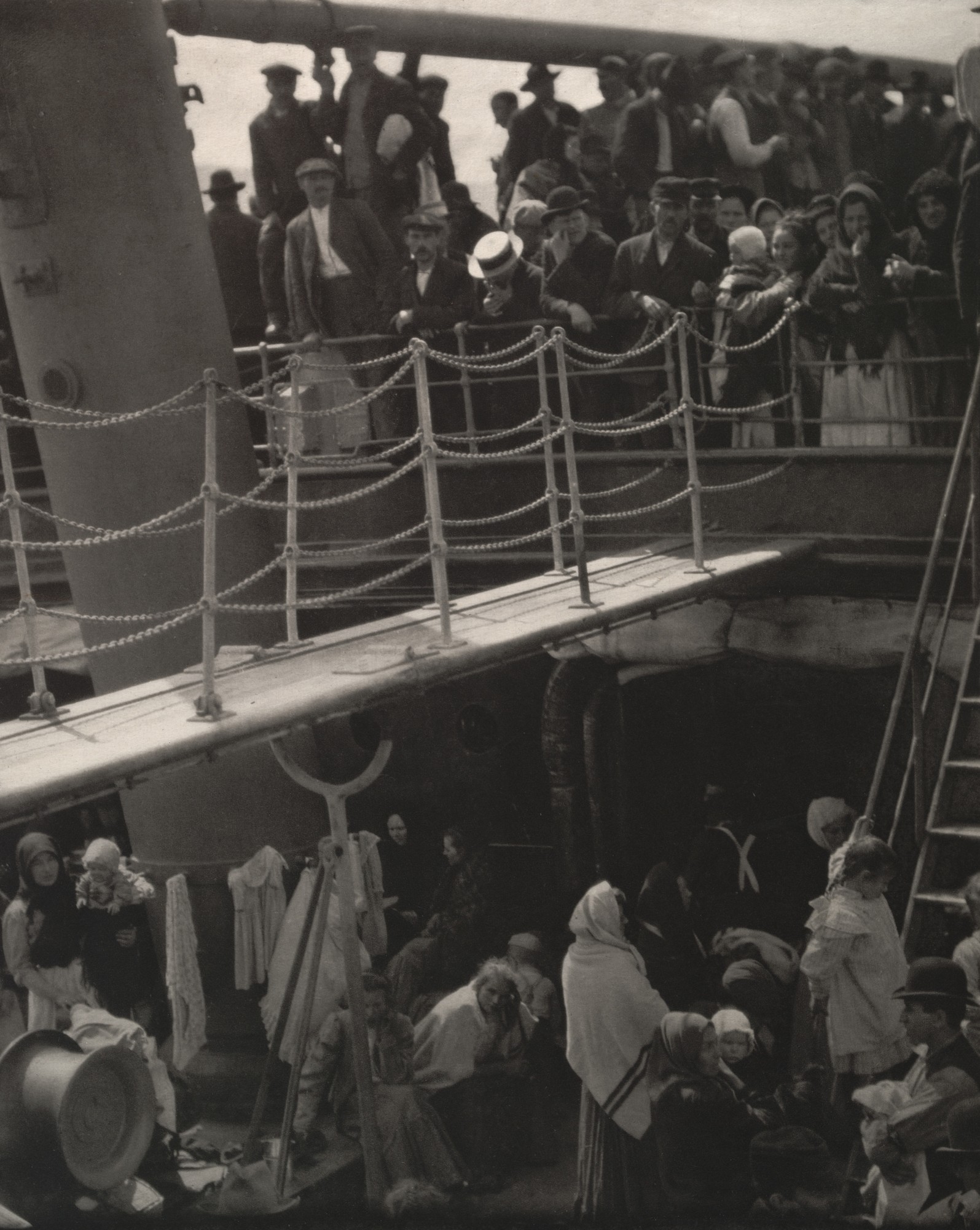 A vertically aligned black and white photo cropped and looking at two decks of a ship. Both the upper and lower decks are full of people. A gangplank leads from the left edge of the photograph to the upper deck.