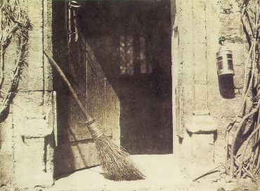 A black and white photo with a warm undertone of an open doorway. A straw broom leans on the door sill.