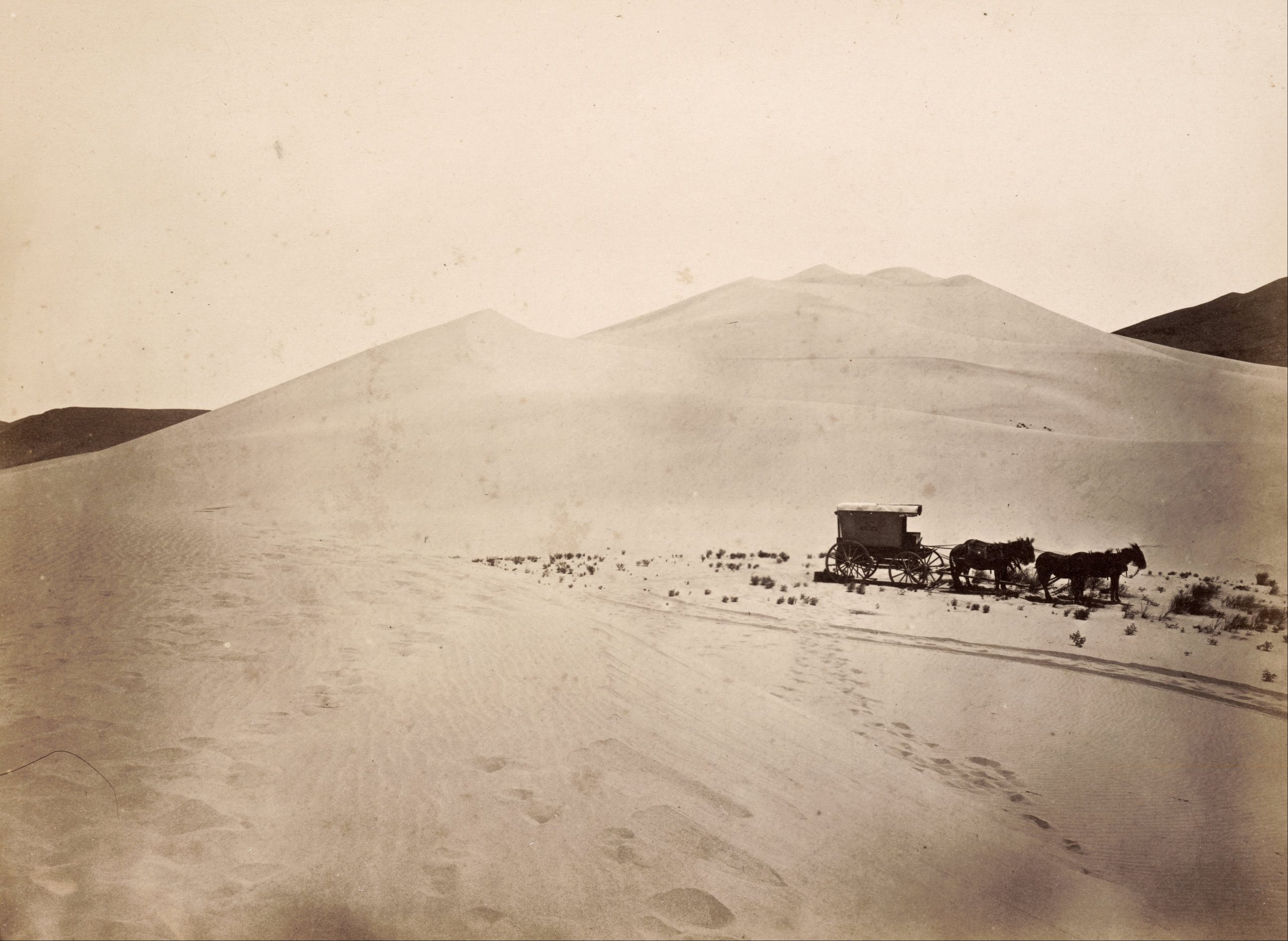 A black and white photograph with a warm undertone of a dessert with hills in the background. A covered cart with horses is in the middle ground on the right side.