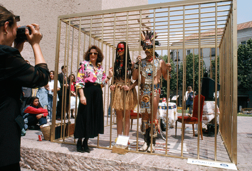 A couple in costume stand in a large gold cage while a woman poses outside of the cage for a photograph with them.