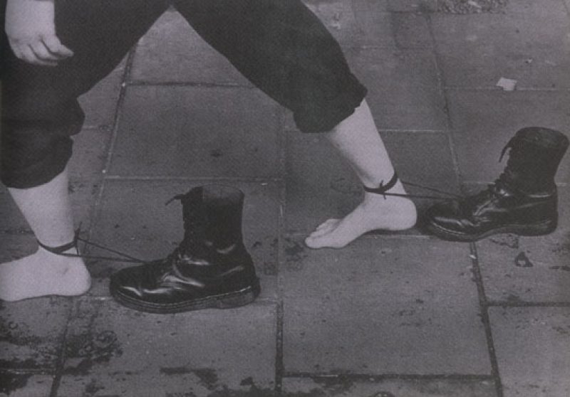 A black and white photograph that shows a pair of legs in dark pants, shown from the knees down. A pair of black combat boots are tied to each ankle, and drag behind the figure, as the figure strides out of the frame.