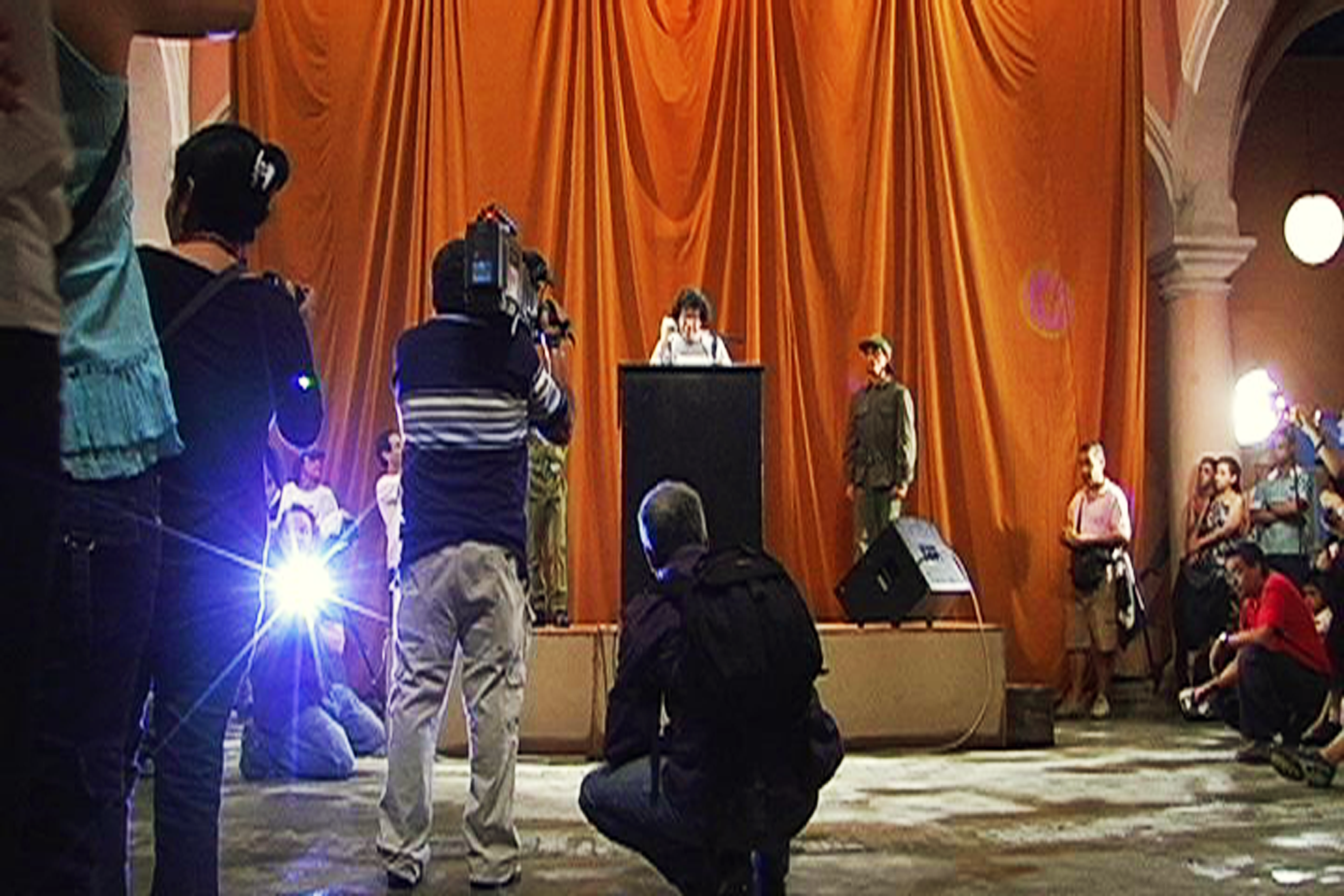 A podium stands in front of an orange curtain, two figures dressed in army fatigues stand on either side. Spectators stand and crouch around the stage, some taking photos and recording.